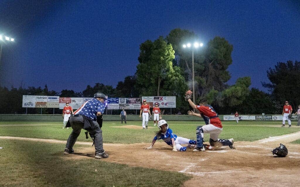 Photo - Madera National’s Leo Barajas slides through the legs of Madera American catcher Carlos Hernandez to score the game-winning run in Tuesday’s District 10 championship game with umpire Scott Varela looking on to make the call. 