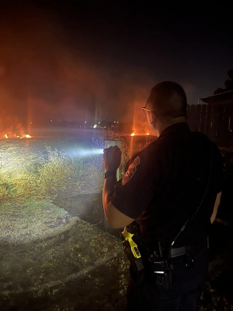 Photo - A Madera Police officer watches a fire in a field to make sure it doesn’t make its way to an apartment complex Thursday evening.