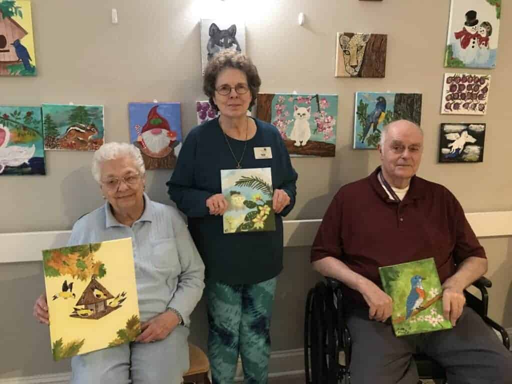 Photo - Betty Robbins, Hazel Toler, and Dale Murphy each show off a picture they painted at Cedar Creek Senior Living. The wall behind them shows paintings created by them and other Cedar Creek residents.