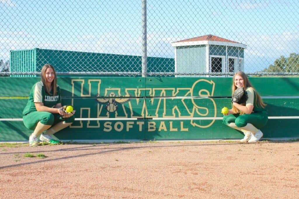 Two softball players kneeling by the Madera Hawks sign on the field.