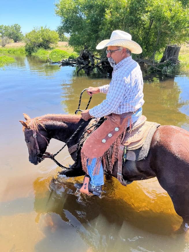 Rancher riding a horse through a shallow water body in a rural setting.