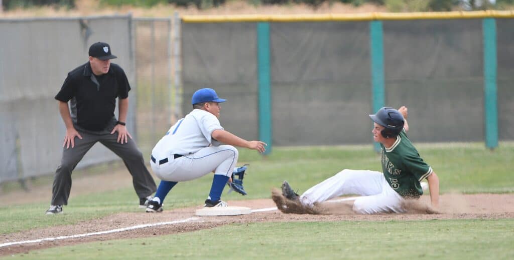 Young baseball player sliding into base during game, with umpire and teammate nearby.