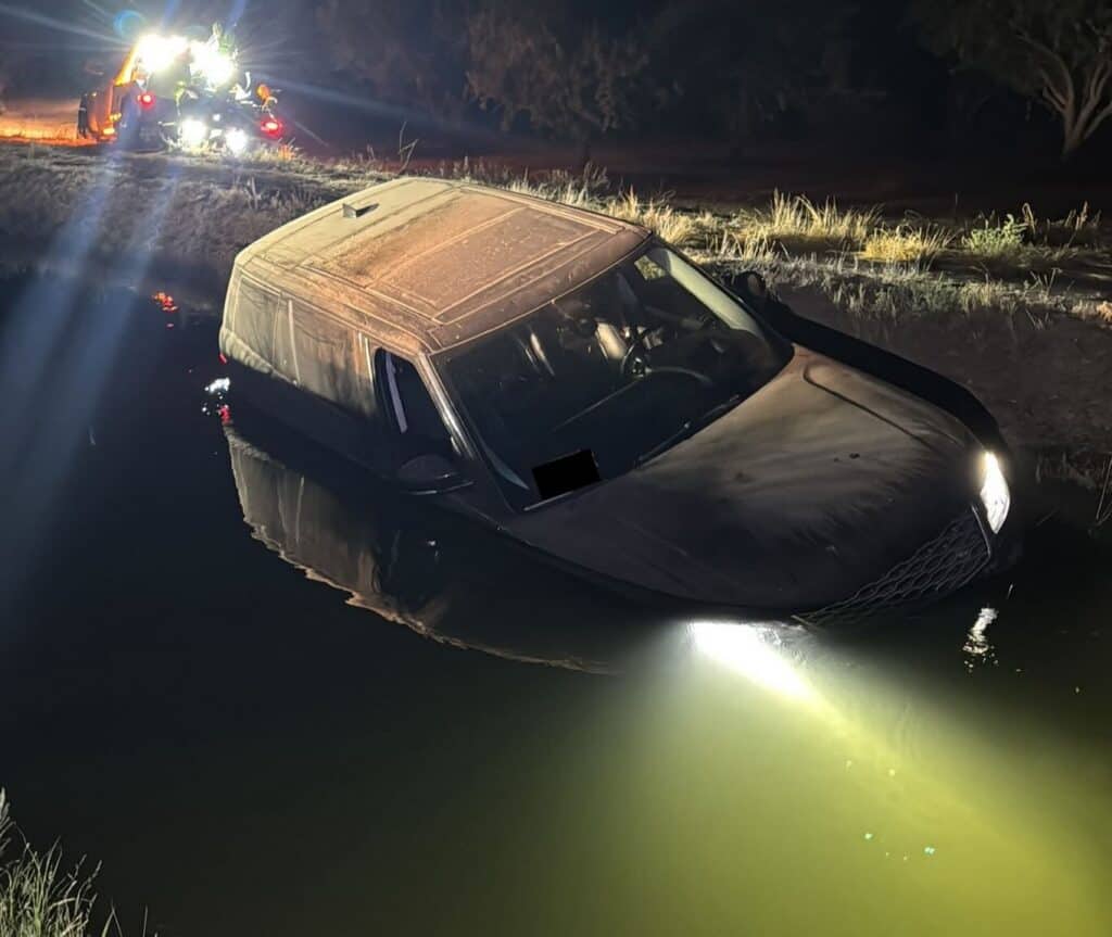 Car partially underwater with headlights on, emergency responders in the background.