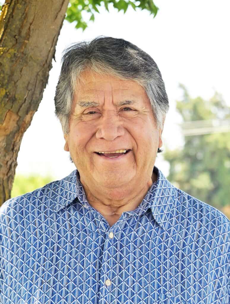 Elderly man smiling outdoors near a tree, wearing a blue patterned shirt, enjoying a sunny day.