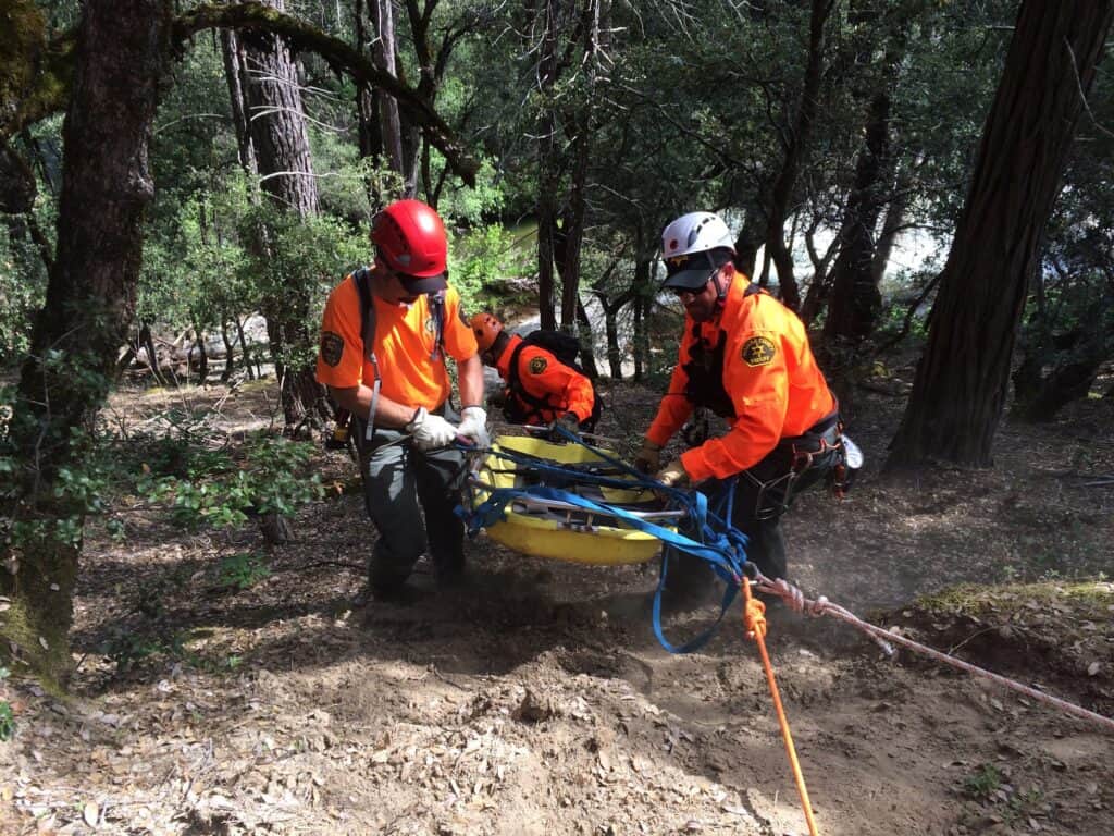 Rescue team members in orange gear and helmets perform a rescue in a wooded area.