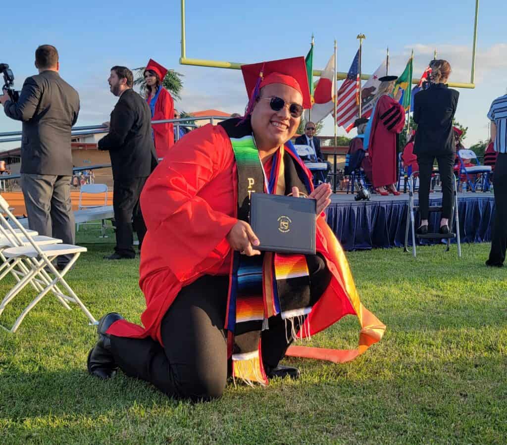 Graduation cap and gown, smiling graduate holding diploma outdoors.