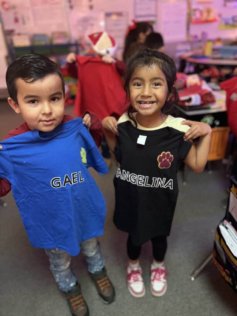 Two children in a classroom holding up personalized T-shirts with names.