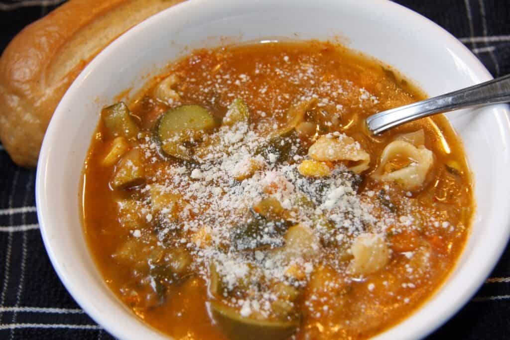 Vegetable soup with zucchini, carrots, and cheese in a white bowl, served with bread.