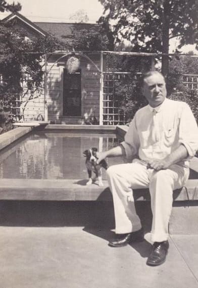 Man sitting by a backyard pool with a small dog, vintage black-and-white photo.