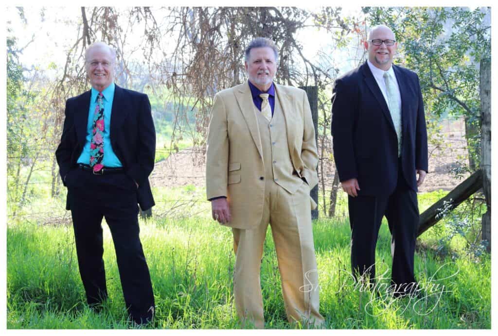 Outdoor photo of three men in suits standing in a grassy field with trees in the background.