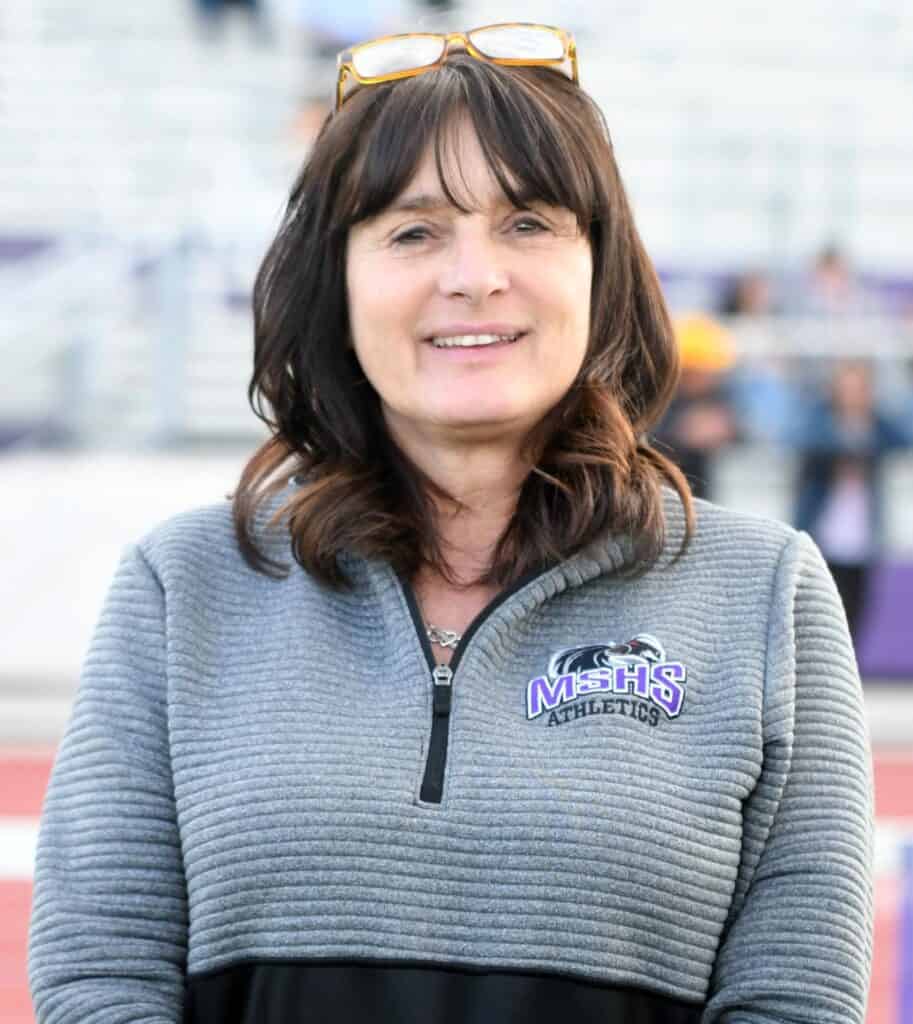 Coach smiling outdoors at track event, wearing MSHS Athletics jacket.