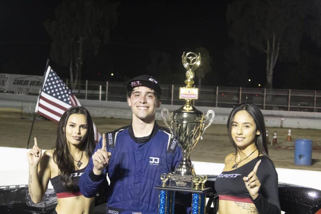 Race car driver celebrating with two women holding an American flag and pointing upward.