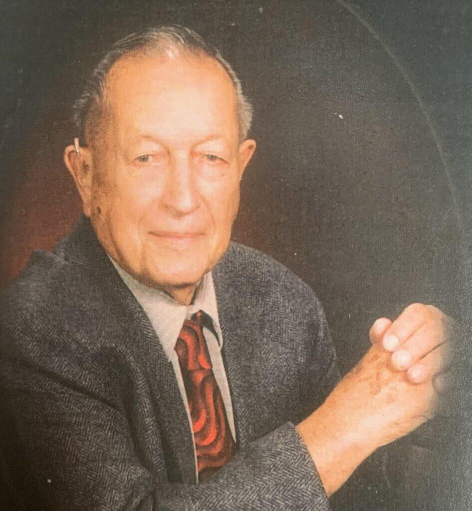 Elderly man in formal attire with a confident expression, sitting with hands clasped.