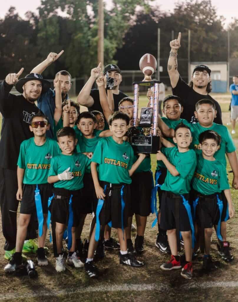 Madera youth football team with trophy and coaches on field after game victory.