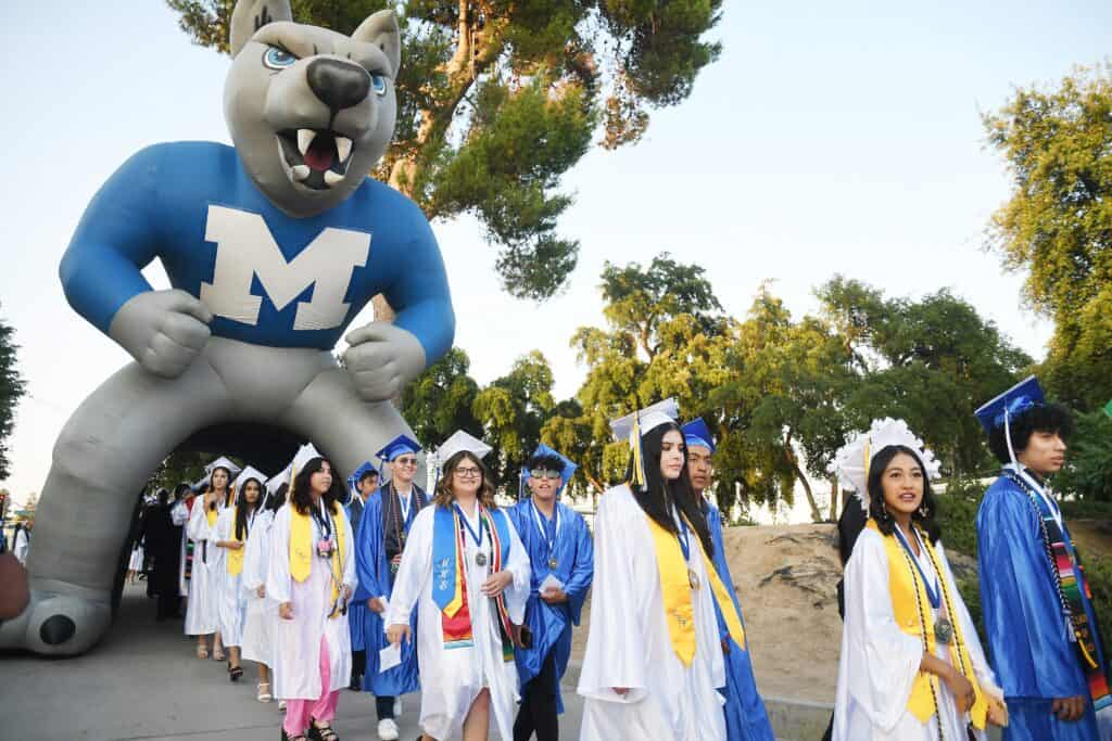 Graduates in blue caps and gowns walking with Madera High mascot in background.