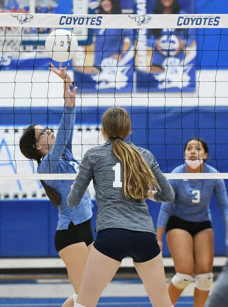Girls playing volleyball at a school gym with "Coyotes" banner.