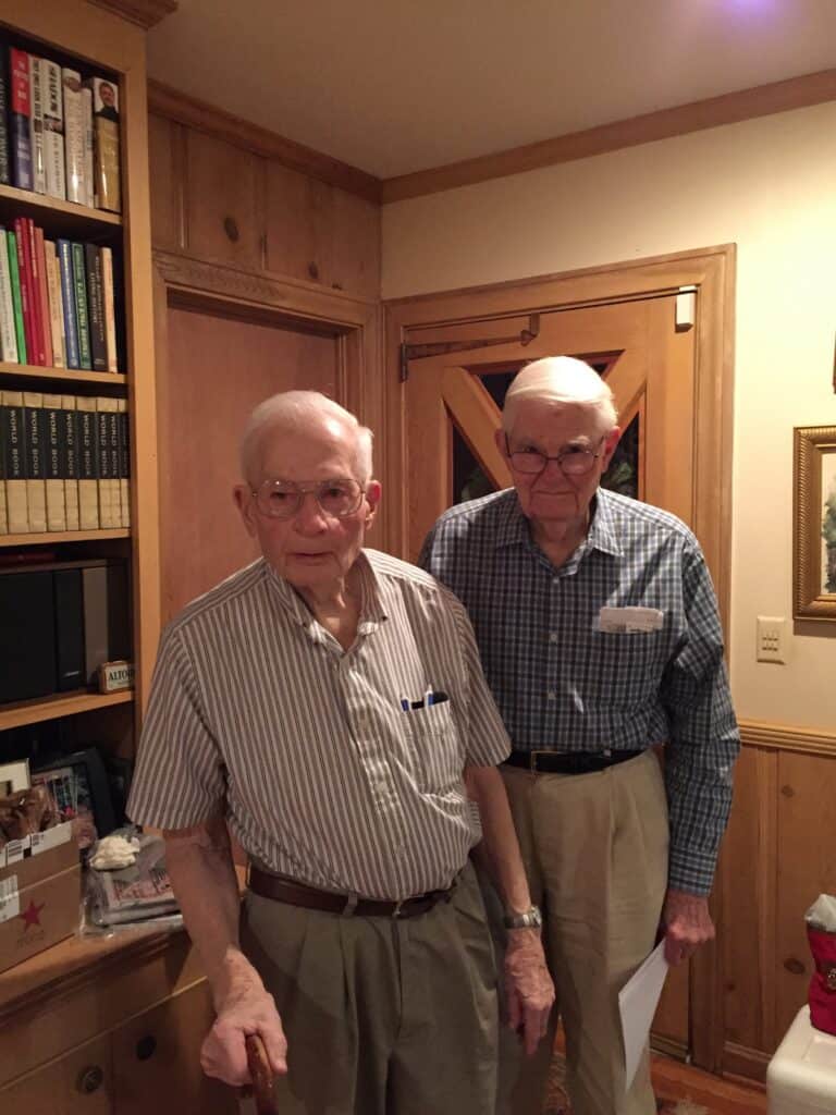 Older men standing together indoors, one with a cane, in a cozy wooden-paneled room.