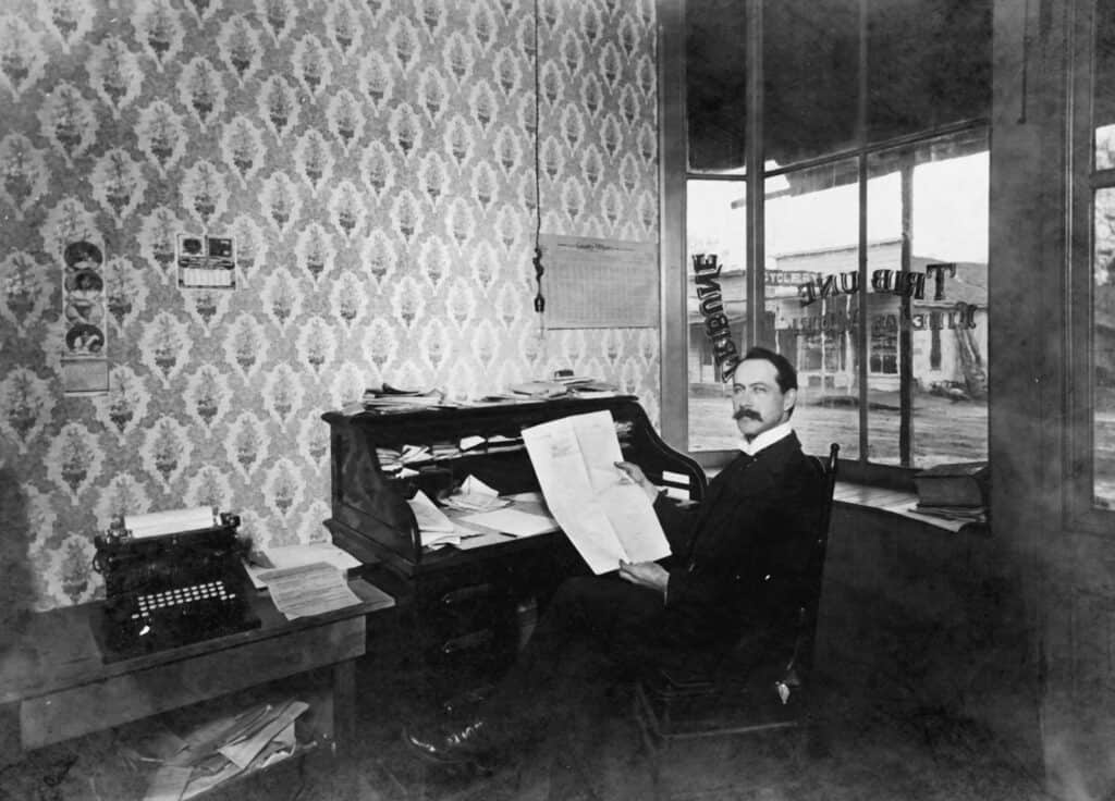 Man working at a vintage desk with papers and typewriter in an office setting.