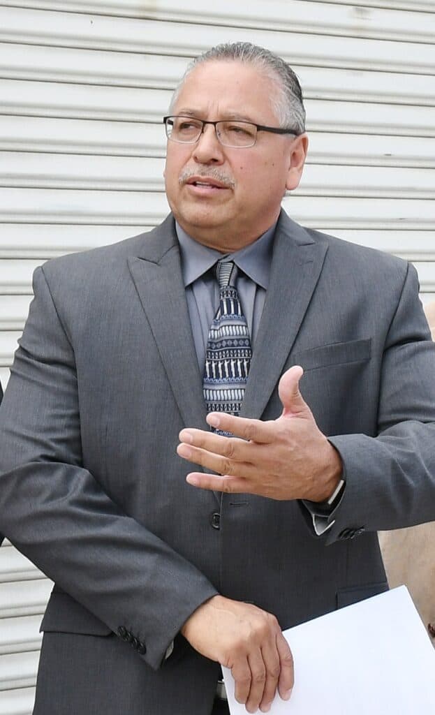Man in suit speaking outdoors, holding papers, in front of a metal shutter.