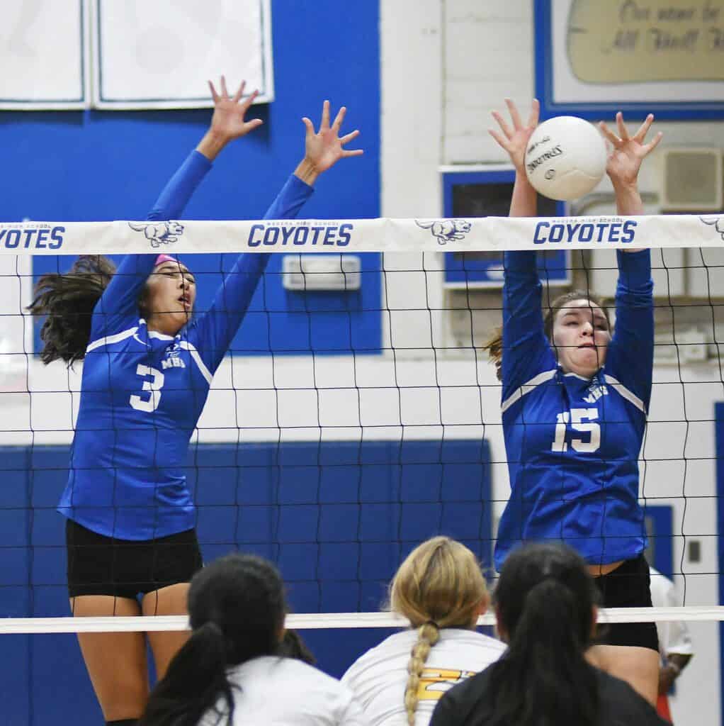 Two female volleyball players in blue jerseys jumping at the net during a match.