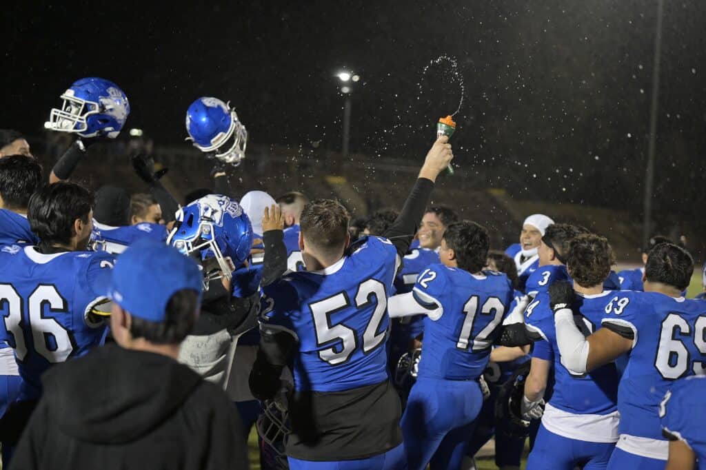 Celebrating victory, Madera High football team in blue uniforms on the field at night.