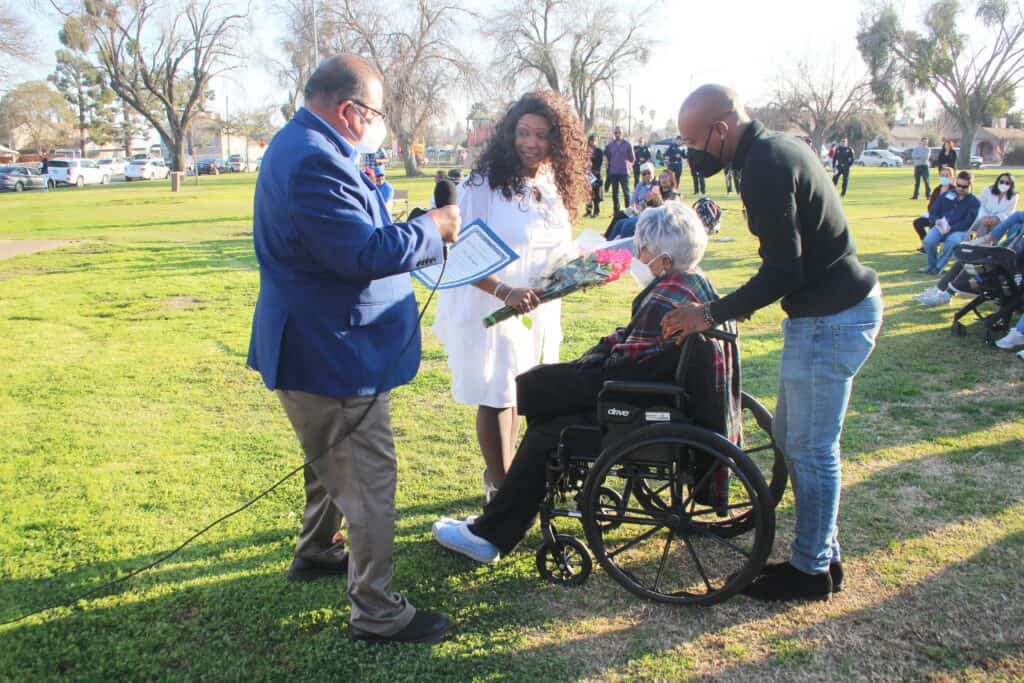 Community gathering with elderly woman in wheelchair receiving flowers.