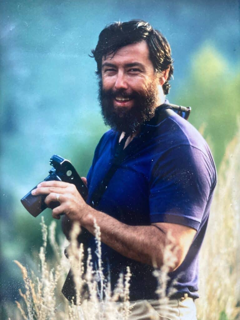 Photographer smiling outdoors holding a camera in a natural setting.