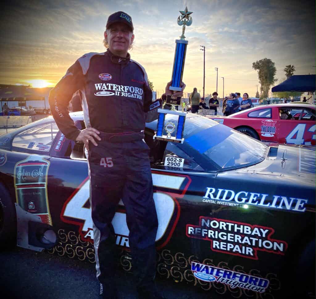 Race car driver holding trophy next to his car at Madera race event.