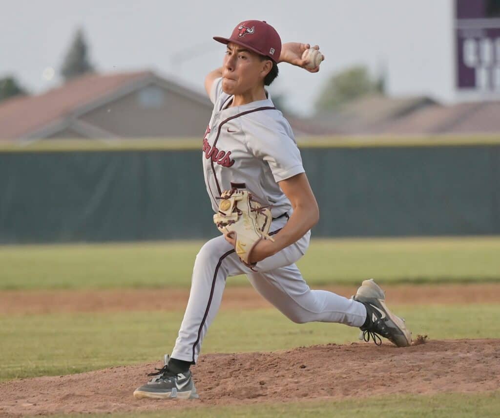 Young baseball player pitching during a game, wearing a maroon cap and uniform, on a sunny day at th.