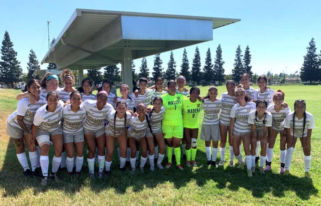 Girls' soccer team posing outdoors with coach and teammates on a sunny day.