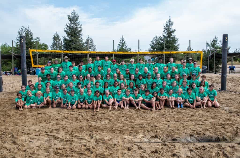 Beach volleyball team posing on sand court with net and trees in background.