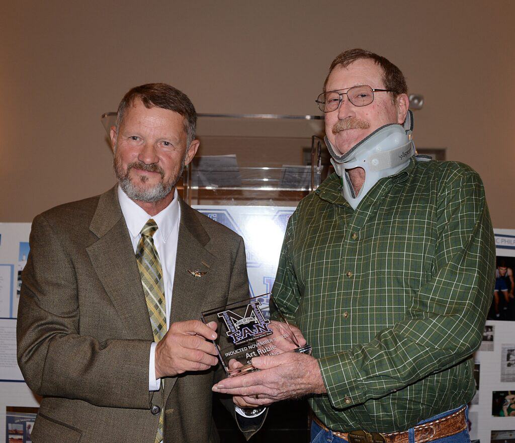 Two men at an award ceremony, one with a neck brace, holding a trophy.