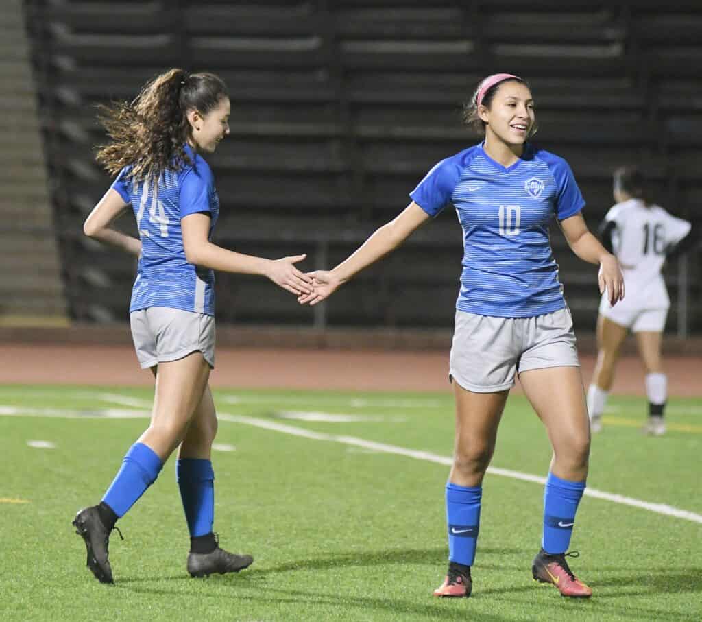 Two female soccer players in blue jerseys celebrating on the field.