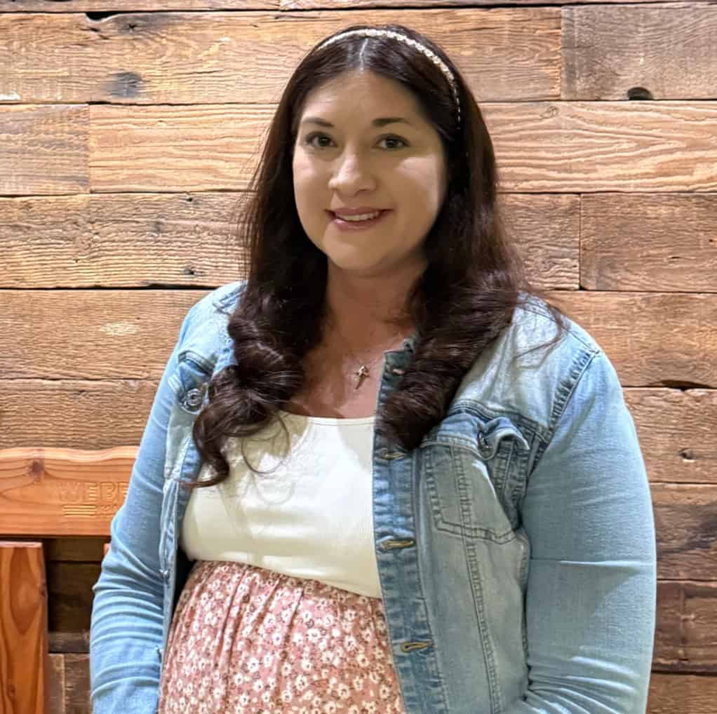 Portrait of a woman smiling in front of a wooden wall, wearing a denim jacket and floral skirt.
