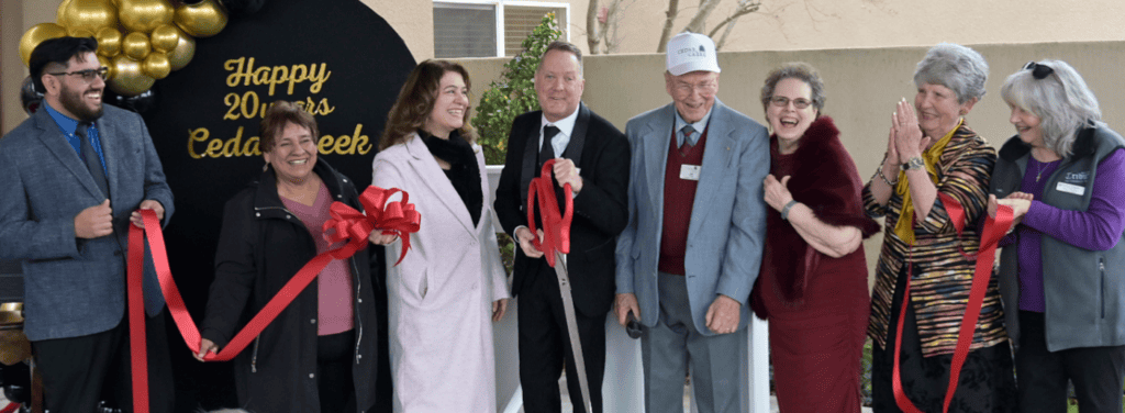 Photo of Cogir of Cedar Creek executive director Rob Huntley, holding scissors, is joined by representatives and presenters for a ribbon-cutting ceremony in honor of Cedar Creek’s 20 year anniversary celebration on January 16.