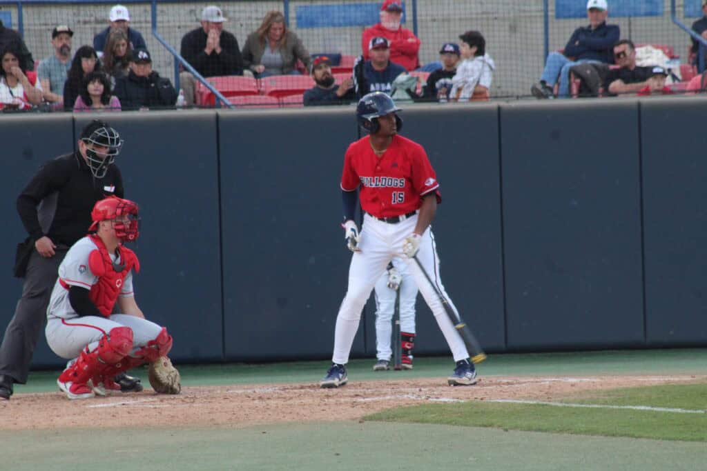 Young baseball player in red uniform at bat during game, with catcher and umpire nearby.