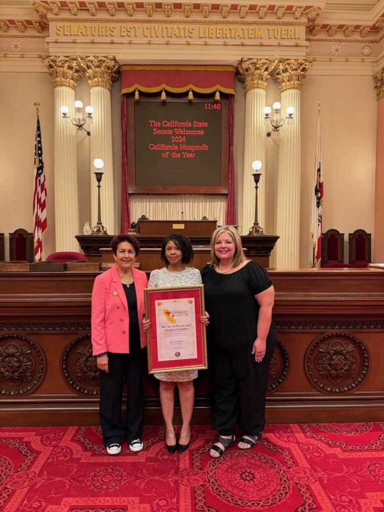 Three women holding a nonprofit award in a historic government chamber.