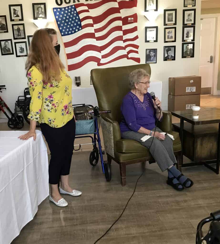 Shirley Sudduth speaking into microphone at event with American flag backdrop.