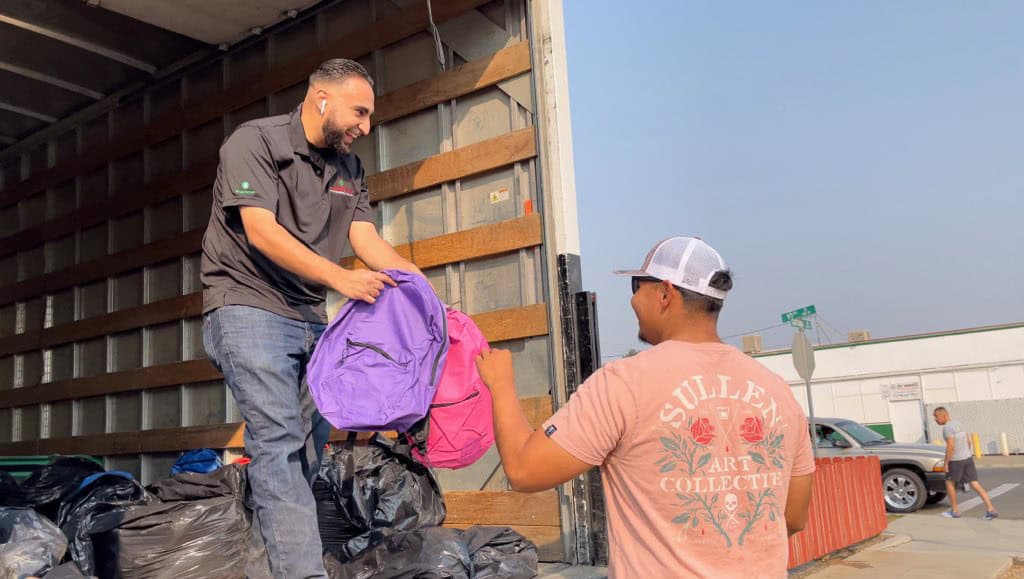 Photo - Pacific Farm Management owner Mike Alamari hands PFM Supervisor Miguel Ortega two of the 750 backpacks his company handed out on Sunday. This is the second year in a row Alamari has held a backpack giveaway.