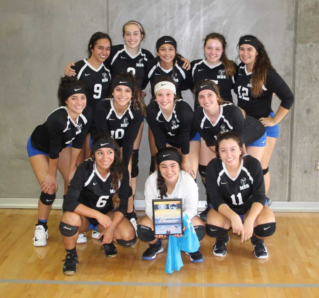 Young female volleyball players in black uniforms pose with a trophy after winning a match.