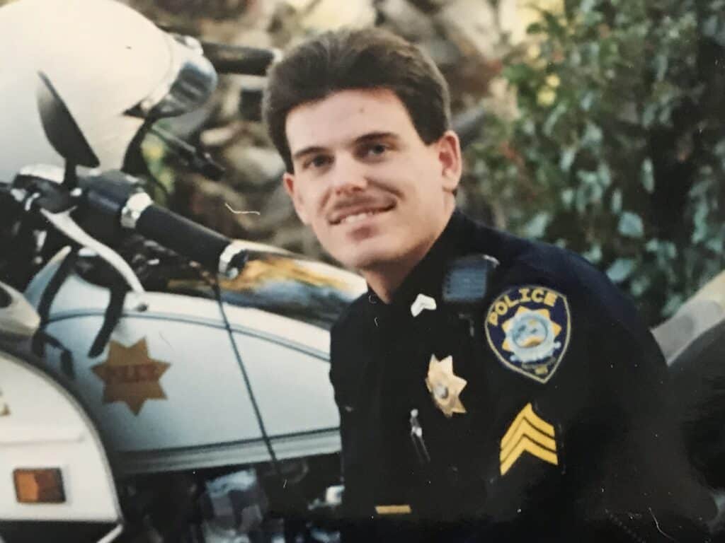 Police officer Kevin Weaver in uniform, smiling, with police vehicle in background.