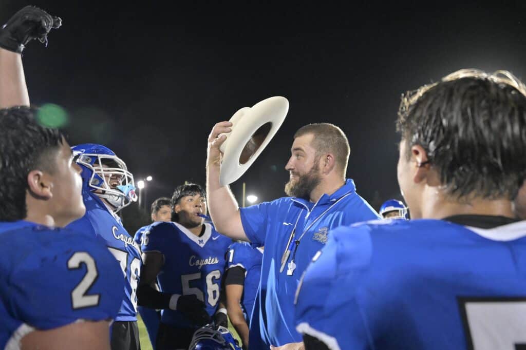 Coyote football coach holding a white hat, surrounded by players in blue jerseys after winning back.