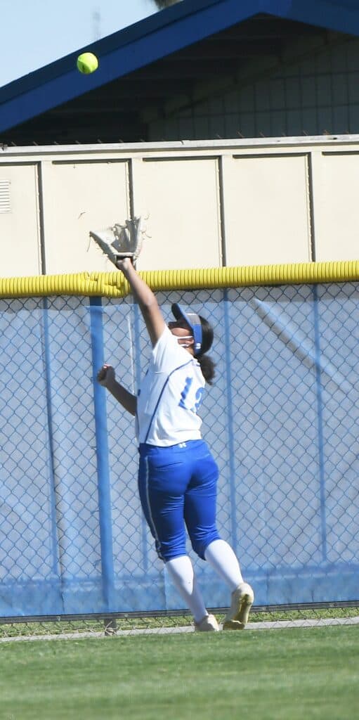 Female softball player jumping to catch a ball near the outfield fence.