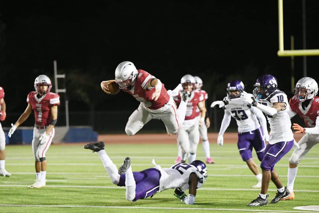 Football player leaps over opponent during game at night.