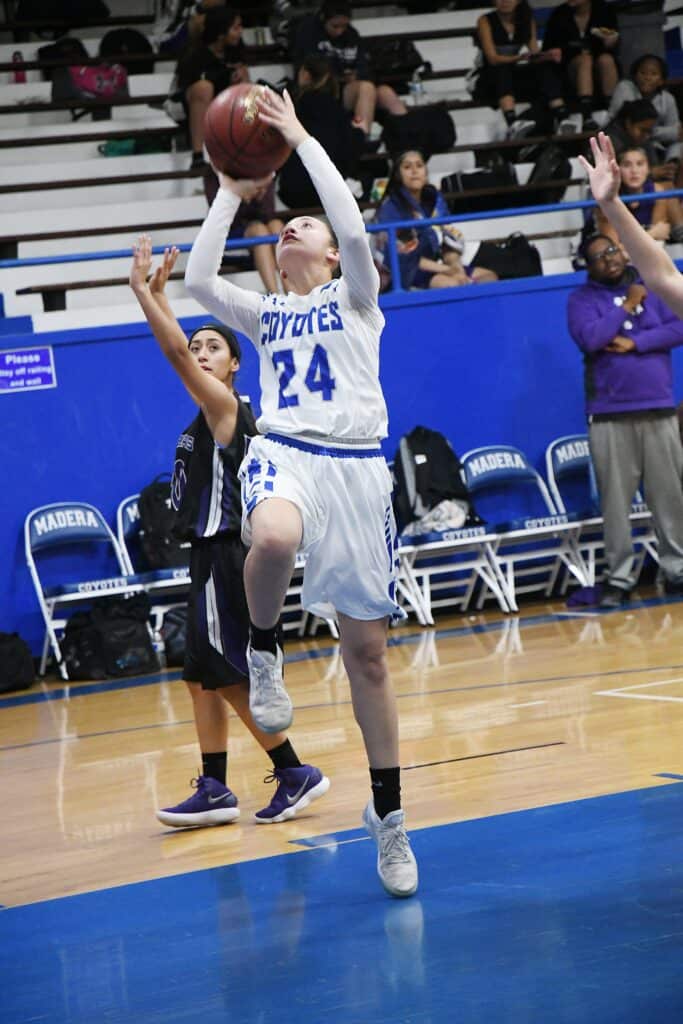 Young female basketball player in white jersey jumping for a shot during game.