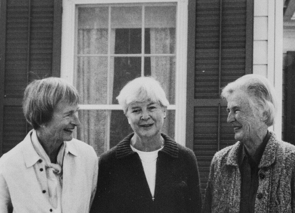 Three elderly women smiling together outside a house.